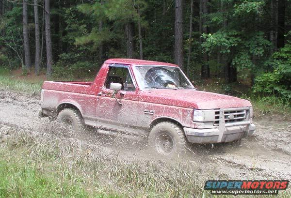 Red pickup truck driving through mud and grass, trees in background