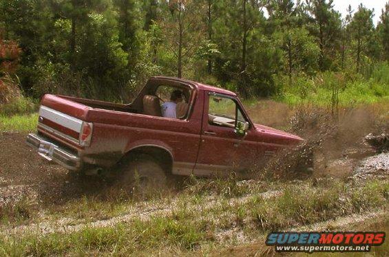 Red Ford Bronco with white stripe on rear quarter panel driving through mud, trees in background