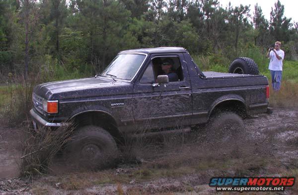Mud-splattered dark blue Ford Bronco driving through mud, spare tire mounted on rear