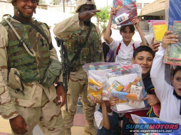 Soldiers in desert camouflage stand with children who are holding bags of school supplies