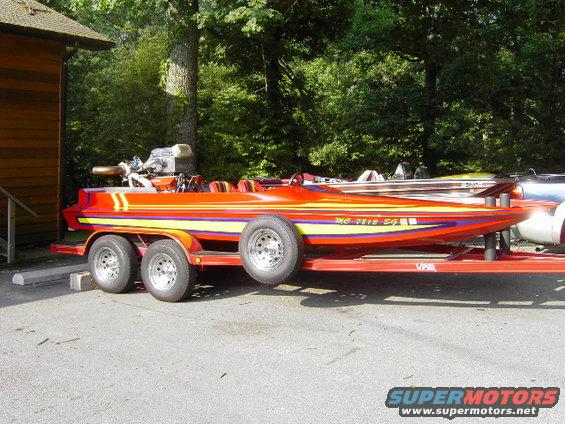 Red speed boat with yellow and purple stripes on a triple-axle trailer, parked near a wooden building. Another boat is visible behind it