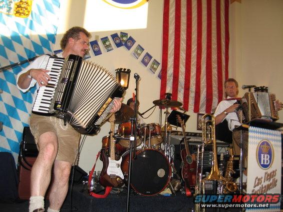 Man in lederhosen plays accordion next to a drum set and another man playing accordion. American flag and Bavarian flags in background