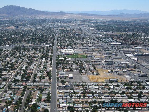 Aerial view of a suburban city with residential neighborhoods, commercial buildings, and a long road, under a clear blue sky with mountains in the distance