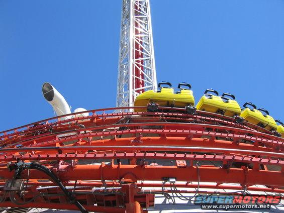 Yellow roller coaster cars on an orange track with a tall white tower in the background under a blue sky