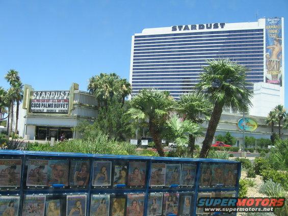 Red car parked in front of Stardust hotel and casino, seen from across a display of magazines
