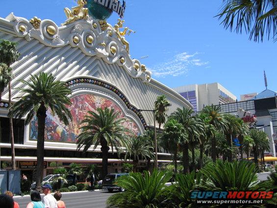 Facade of a large building with ornate gold trim and a mural, surrounded by palm trees under a blue sky. A street with cars and people is in the foreground
