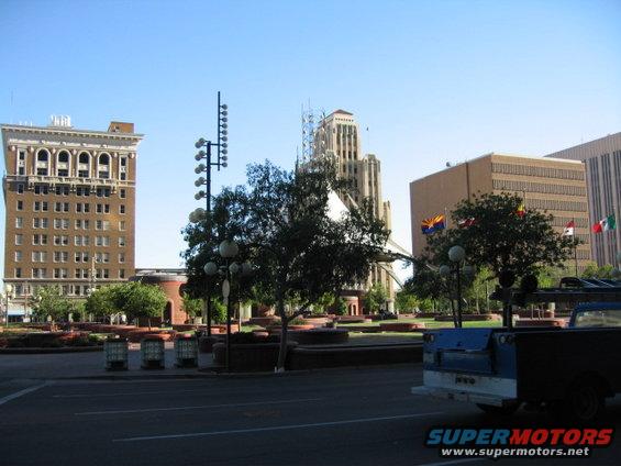 Blue pickup truck parked in a city square with buildings and trees. SUPERMOTORS.NET logo visible