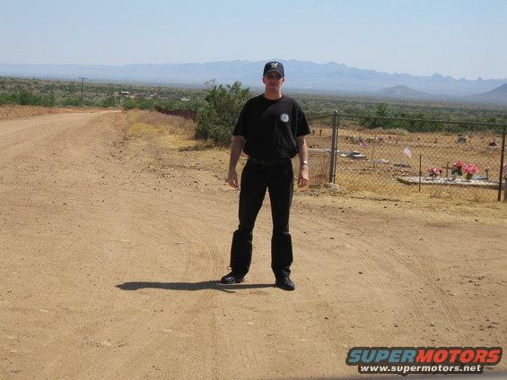 Person in black t-shirt and pants stands on a dirt road in a dry, arid landscape with mountains in the distance