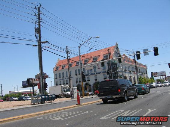 Black SUV driving on a street in front of a building with a red roof. Other cars and a truck are visible