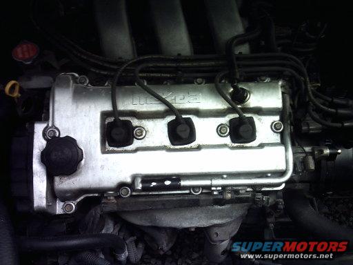 Engine bay with silver valve cover showing "Mazda" text, black spark plug wires, and hoses