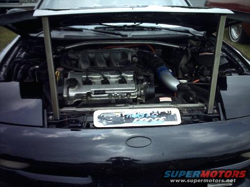 car engine bay with hood open, showing engine, intake, and some wiring. A small graphic of a car is on a plate in front of the engine