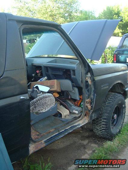 Side view of a beat-up Ford Bronco with the door open and hood up. Interior is messy, with a leopard print seat cover