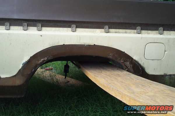 Side view of a vehicle's rear wheel well and fuel door. A wooden plank is positioned under the wheel well
