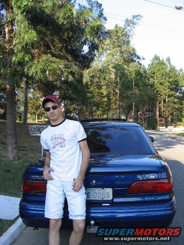 Man in white t-shirt and shorts standing by a blue 1995 Ford Taurus. License plate reads E-0098