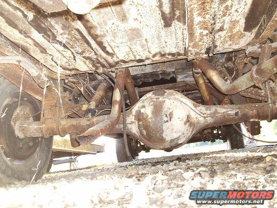 Underside of a heavily rusted truck, showing the rear differential, exhaust pipes, and frame. The vehicle appears to be in poor condition