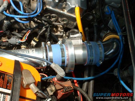 Engine bay showing an aftermarket intake tube with blue couplers and clamps, connected to a throttle body. A yellow battery is visible