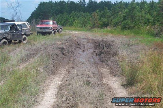 Dark Bronco and red pickup truck on a muddy path with trees in background