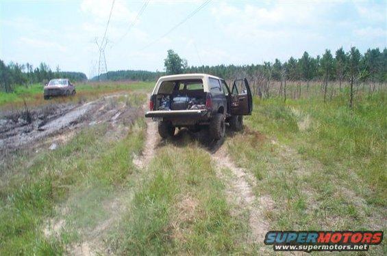 Dark brown Ford Bronco with a white top, rear doors open, parked on a dirt track next to a muddy area