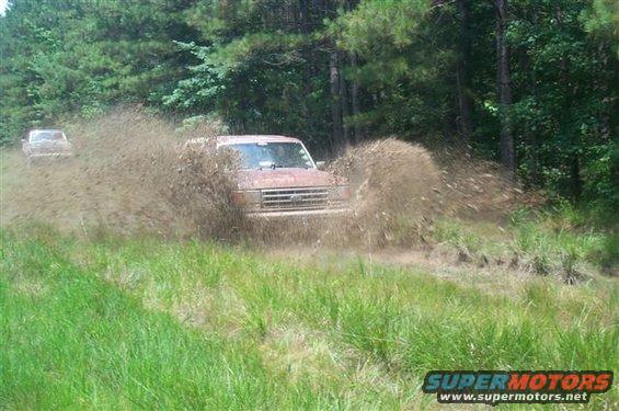 Ford Bronco driving through mud, kicking up a lot of spray. Another vehicle is visible in the background