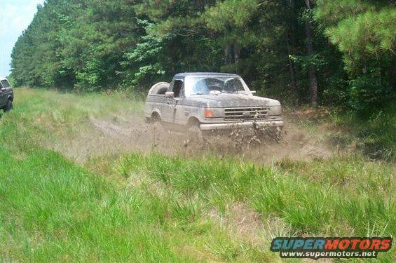 Mud-splattered 1987 Ford Bronco driving through a muddy field with trees in the background