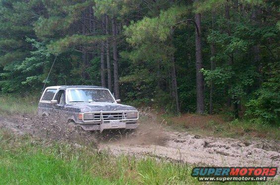 Dark 1987 Ford Bronco driving through mud on a wooded trail