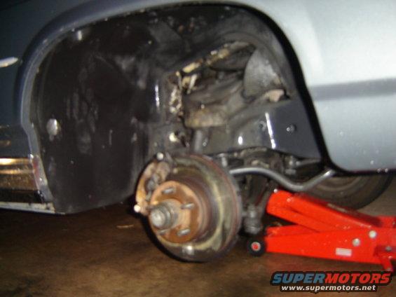 Close-up of a car's front wheel well, suspension components, and a red hydraulic jack supporting the vehicle