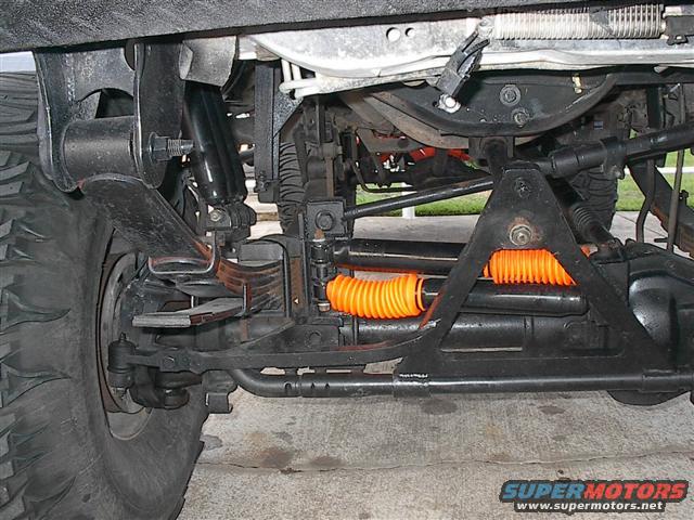Underside view of a truck's front suspension, showing leaf springs, shocks, and steering components with orange bellows