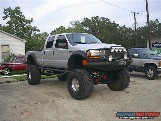 Lifted silver four-door pickup truck with large tires and a brush guard. Another pickup truck is parked behind it