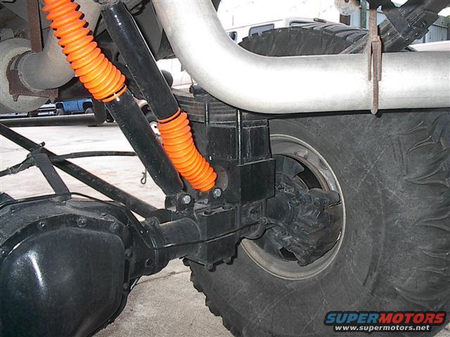 Rear suspension with orange shock boots, large tire, and exhaust pipe. Another vehicle is visible in the background