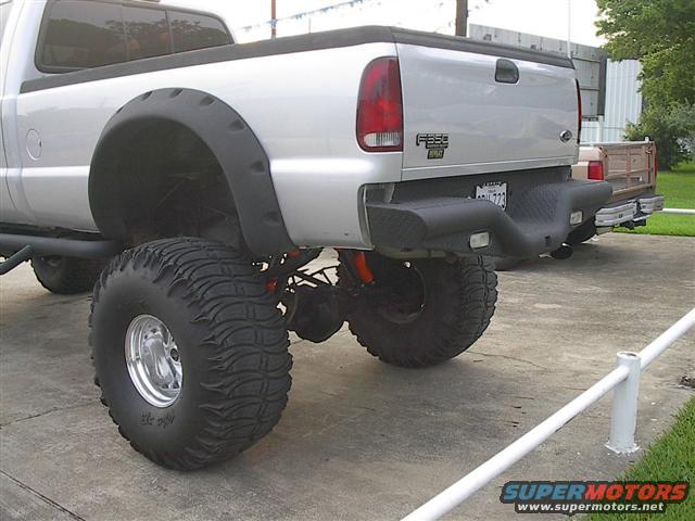 Silver pickup truck with large tires and fender flares, rear view. A second truck is partially visible behind it