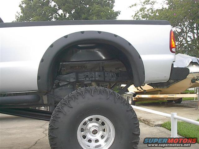 Silver pickup truck's large rear tire and wheel well. A boat on a trailer is visible behind it