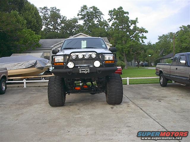 Lifted pickup truck with big tires and extra lights on front, parked next to a boat on a trailer and another pickup truck