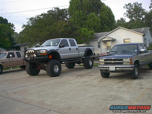 Two lifted pickup trucks parked in a driveway, one silver and one blue. A third truck is partially visible on the left