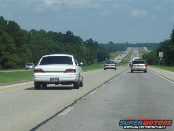 White sedan driving on a highway, other cars in distance. License plate is visible