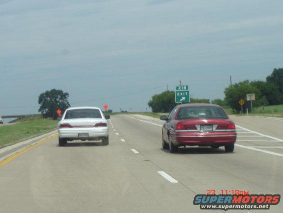 Red and white sedans driving on a highway under a cloudy sky. Exit 218 sign visible