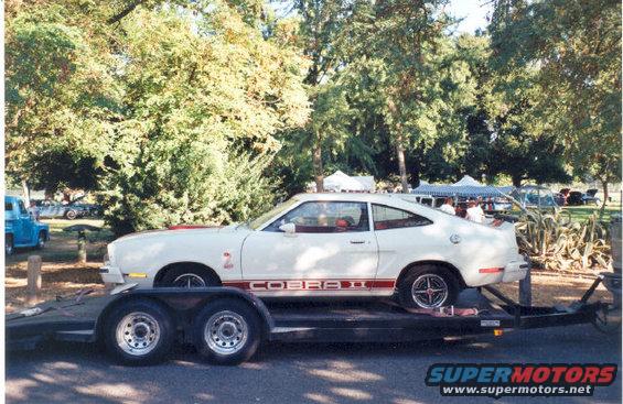 White 1977 Ford Mustang II Cobra II on a trailer, side view. Red stripe and 'COBRA II' text on the door. Red '2' badge on fender