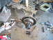 1995 Ford Bronco - Front suspension and brake components are disassembled, with tools like a C-clamp, screwdriver, and hammer nearby on the concrete floor.