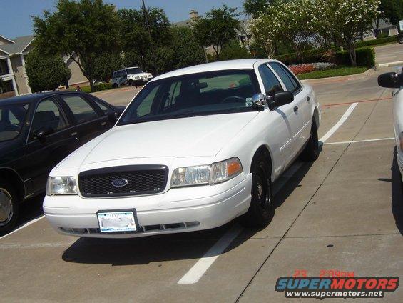 White Ford Crown Victoria parked in a lot, front view. Another dark car is partially visible to the left
