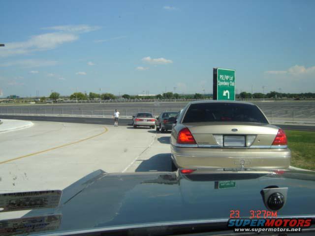 Rear of a gold Ford Crown Victoria, parked on a road near a speedway. Another car is behind it
