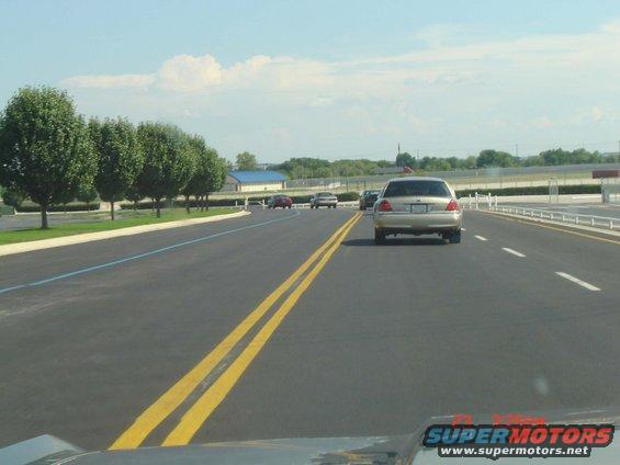 Silver Ford Crown Victoria driving on a highway, rear view. Visible text 'SUPERMOTORS'