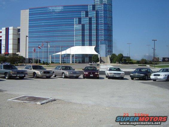 Several Ford Crown Victorias parked in front of a modern building, one burgundy, others silver and white