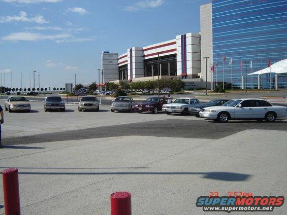 Several sedans parked in a lot with a large building in the background. The white sedan on the right appears to be a 1998 Ford Crown Victoria