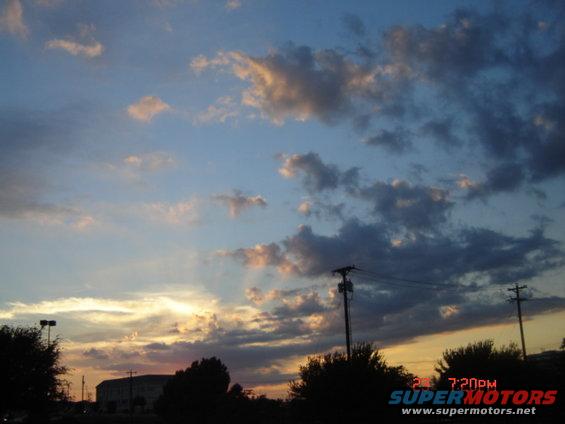 Sunset sky with clouds, utility poles, and silhouetted trees and buildings. Watermark reads SUPERMOTORS.NET