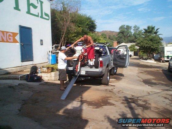 Two guys loading a dirt bike into a pickup truck next to a building