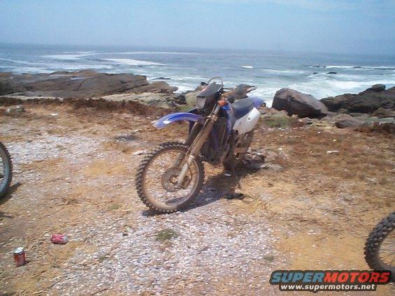Two dirt bikes parked on a rocky shore with the ocean in the background. Cans are on the ground