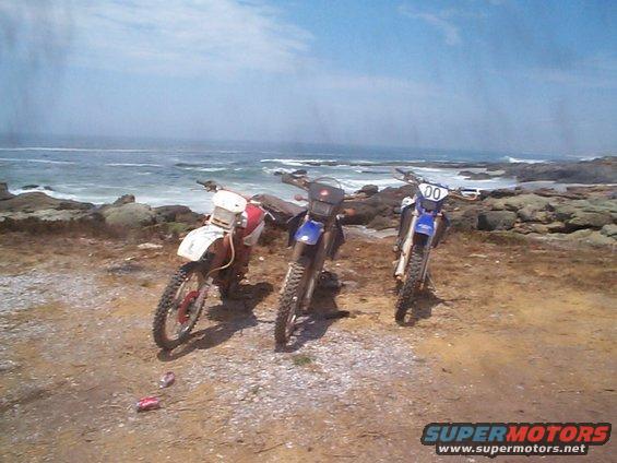 Three dirt bikes parked on a rocky shore with the ocean in the background
