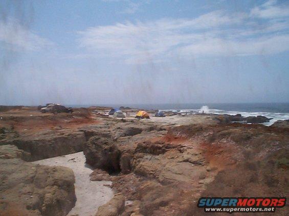 Tents set up on rocky shore with vehicles parked nearby, ocean in background