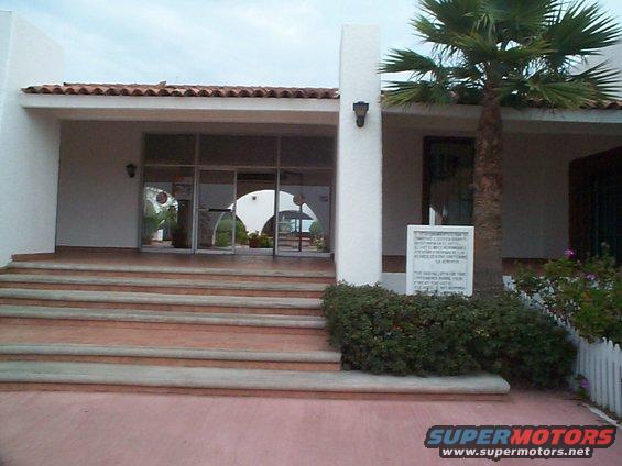 Building entrance with tiled roof, glass doors, and a sign next to a palm tree. Supermotors logo at bottom right