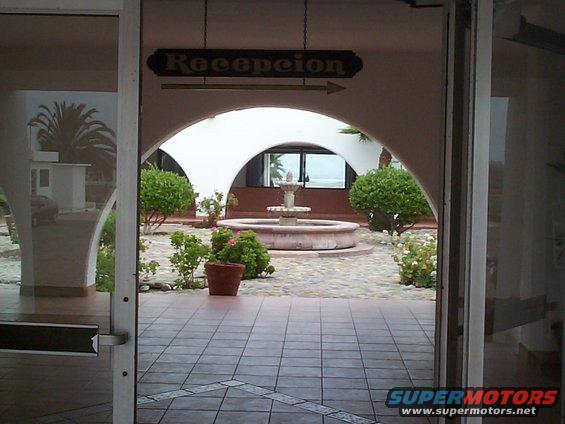 View through doorway to a courtyard with a fountain, plants, and a sign that says 'Recepcion'