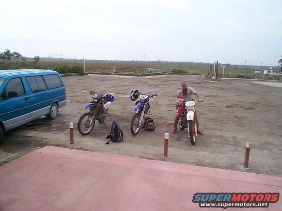 Blue van parked next to three dirt bikes, one with a rider, on a dirt lot